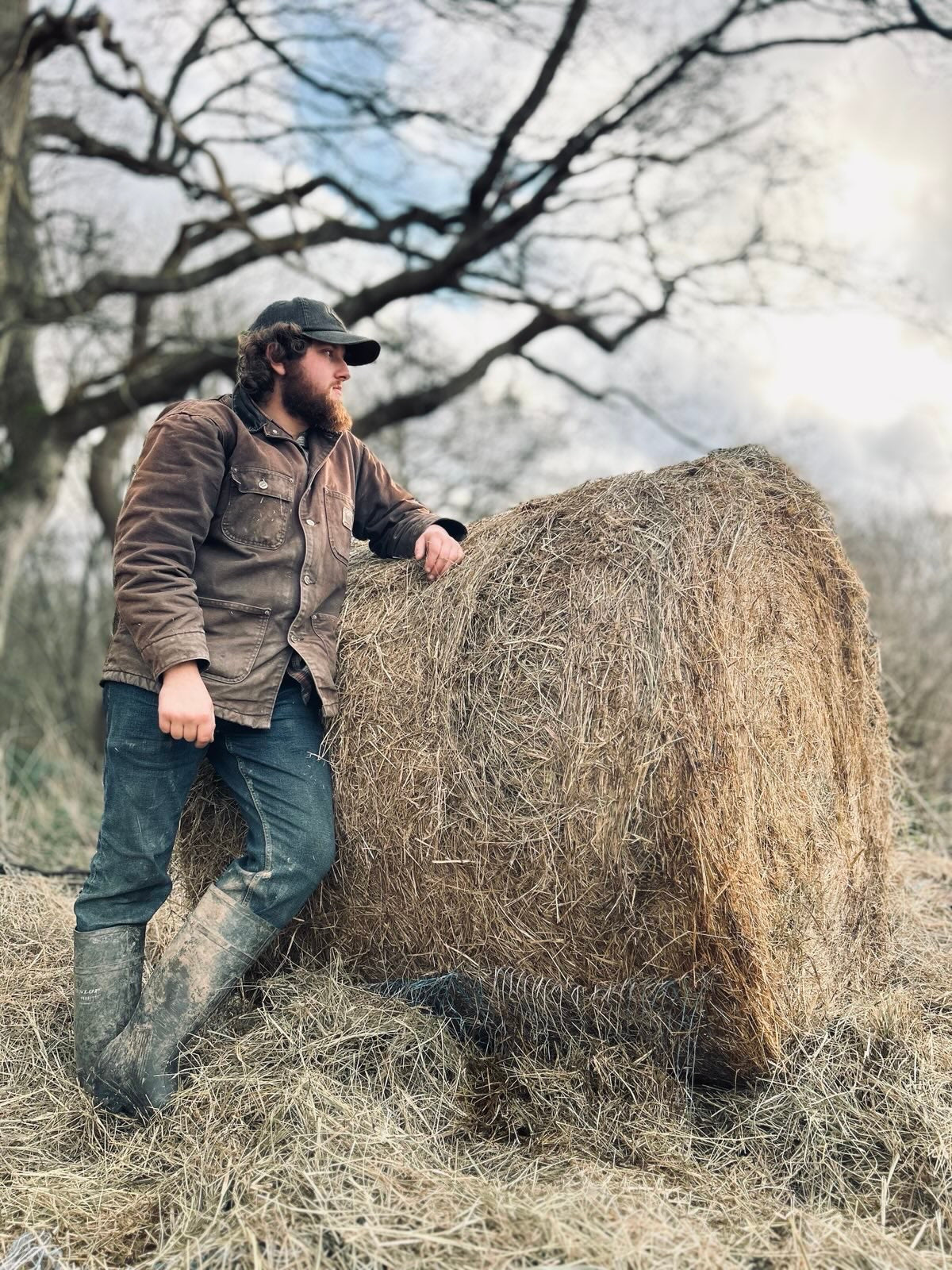 Man leaning against a large hay bale in an outdoor setting with trees and open sky.